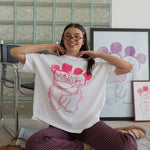 Person sitting on the floor wearing a white t-shirt with a graphic design, surrounded by framed artwork in a room with a glass block wall.