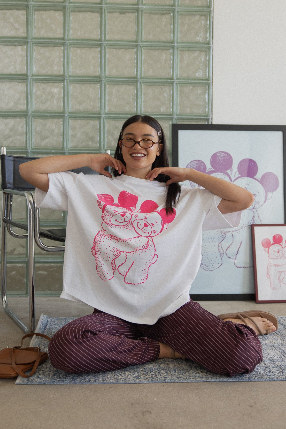 Person sitting on the floor wearing a white t-shirt with a graphic design, surrounded by framed artwork in a room with a glass block wall.