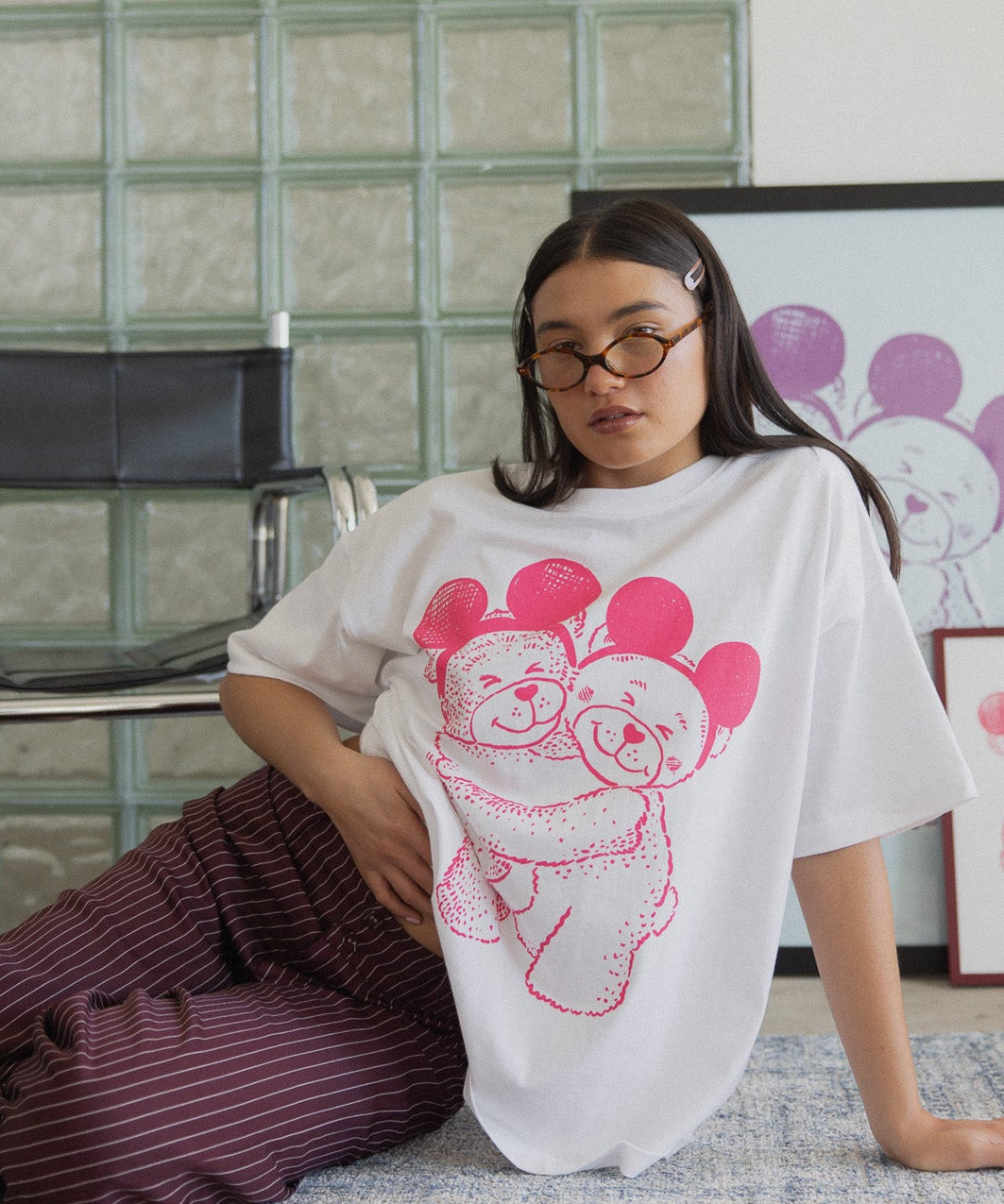 Person wearing a white t-shirt with pink graphic design sitting on the floor against a glass block wall.