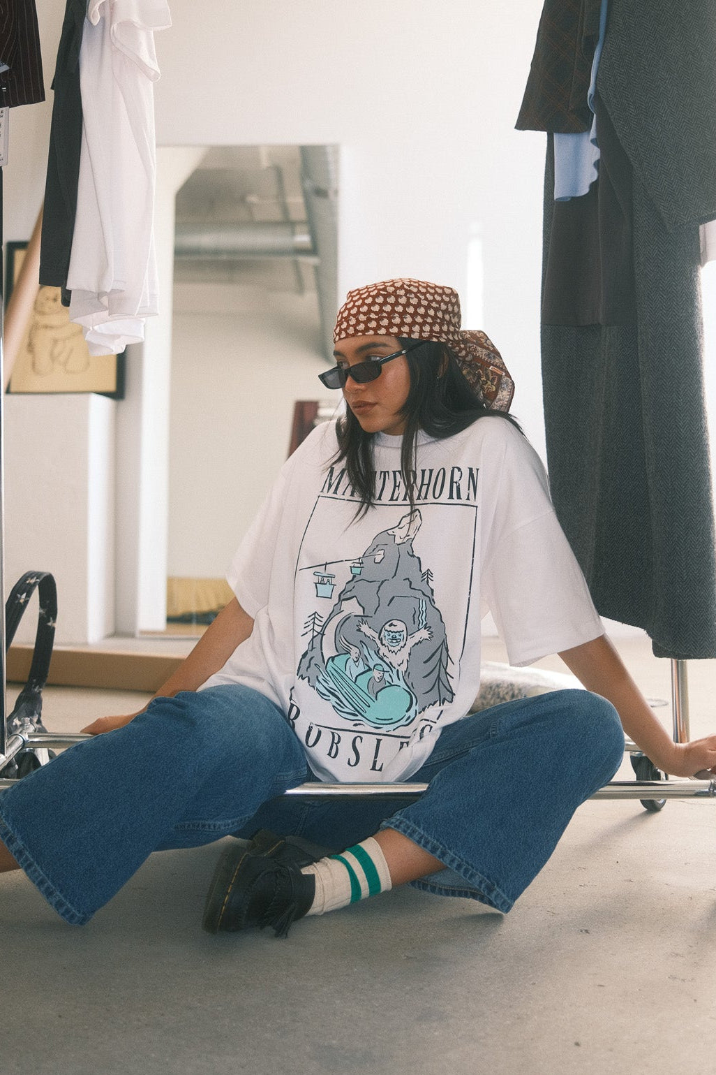 Person sitting on a chair wearing a graphic t-shirt and sunglasses in a clothing store.