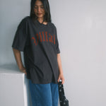 Person wearing a black oversized t-shirt with red text and blue pants, holding a black suitcase against a plain background.