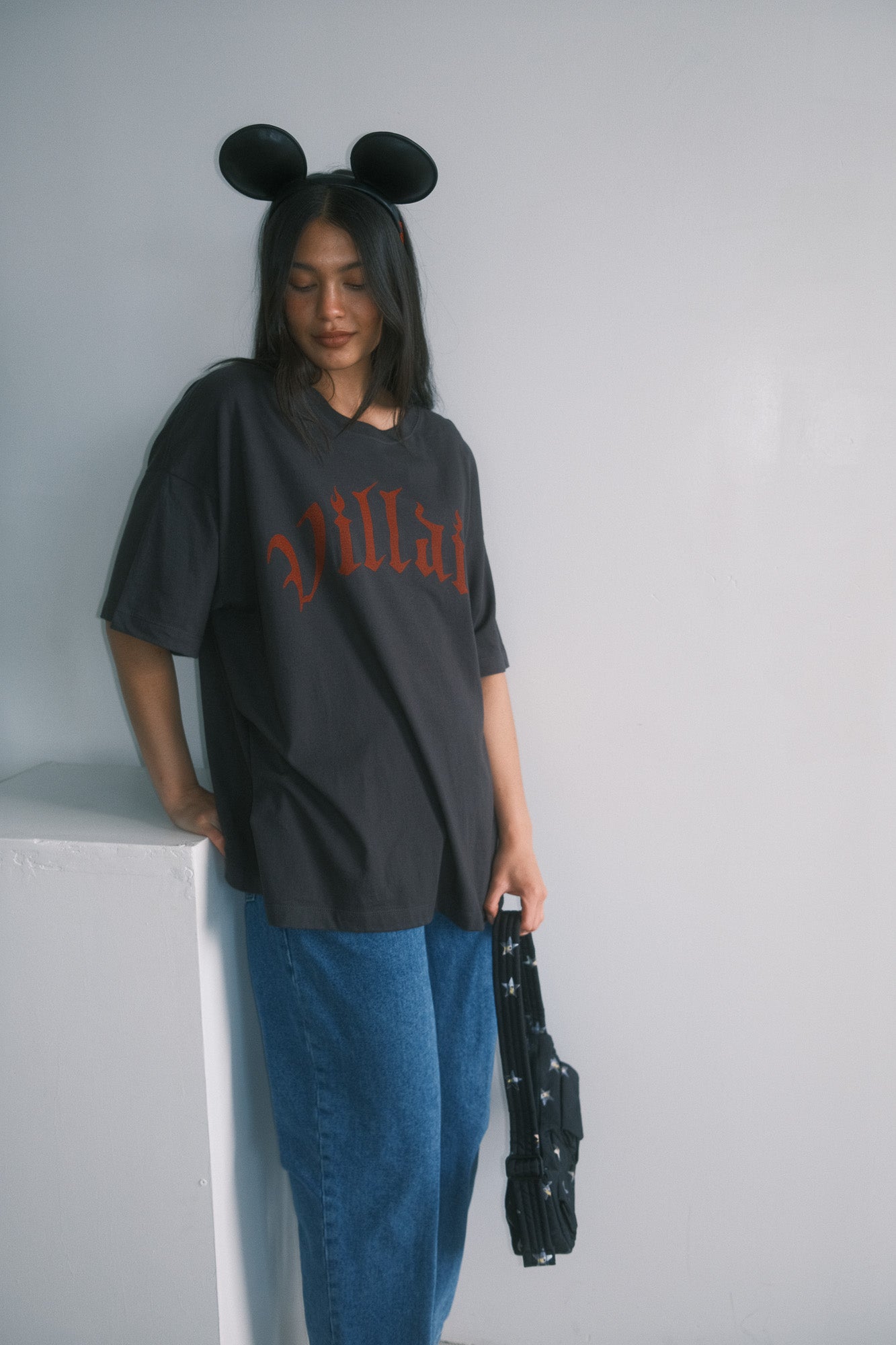 Person wearing a black oversized t-shirt with red text and blue pants, holding a black suitcase against a plain background.